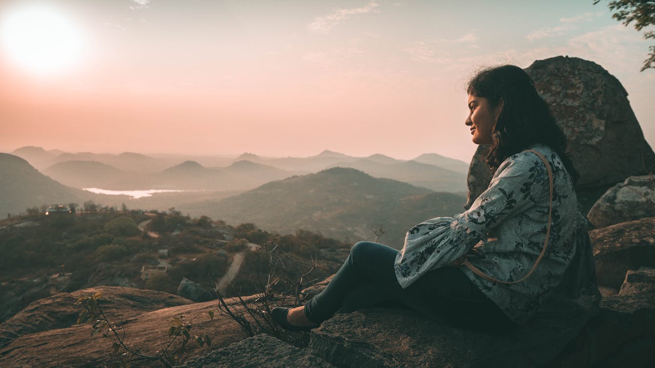 Woman sitting on a rock admires a breathtaking sunset over mountain landscape.