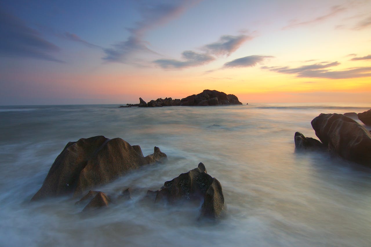 portfolio-img-03 A serene seascape at sunset with rocks and gentle waves under a colorful sky.